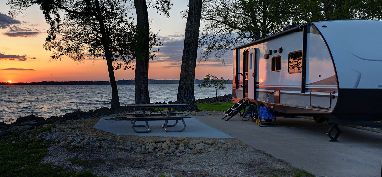 An RV and picnic table on the shore of a lake at sunset