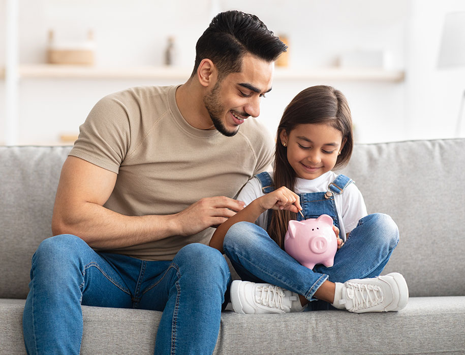 Dad helping daughter put money in piggy bank at home on couch